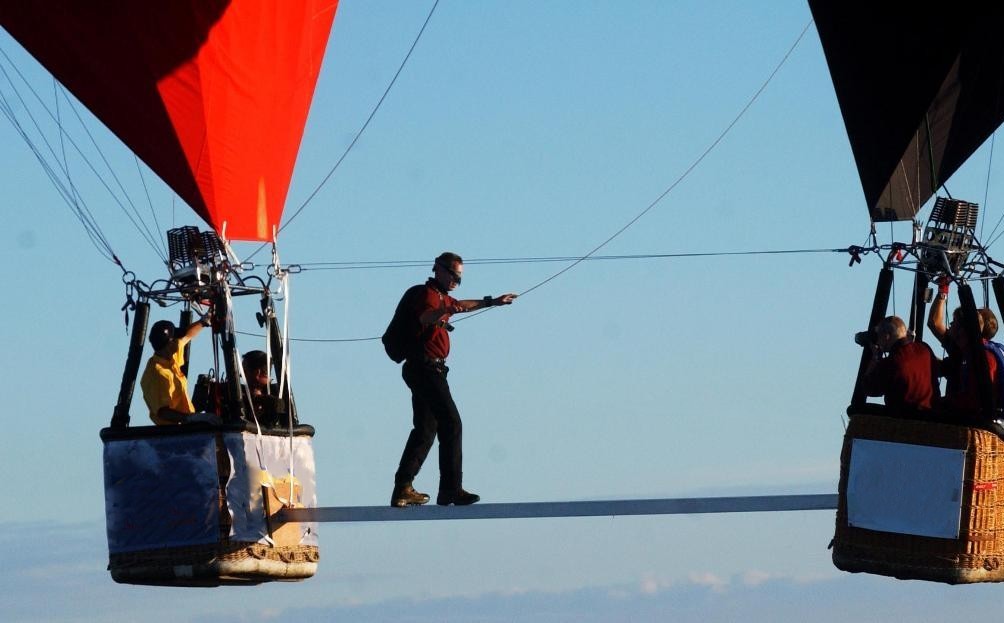 The image is of the tightrope walk, it shows the scoop and basket of both balloons, the aluminium pole between them, and steel wires linking the burners frames, with Mike part way through his walk.